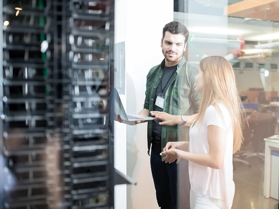 Ein Mann mit Lanyard um den Hals erklärt einer Frau am Laptop etwas in einem Serverraum 